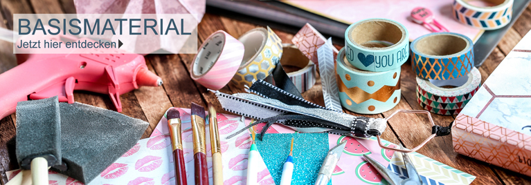 Verschiedenes Bastel-Basismaterial auf einem Holztisch, darunter Hei�klebepistole, Schwammpinsel, Pinsel, Washi-Tapes, Glitzerpapier, Cutter und Deko-B�nder. Text im Bild: BASISMATERIAL - Jetzt hier entdecken.